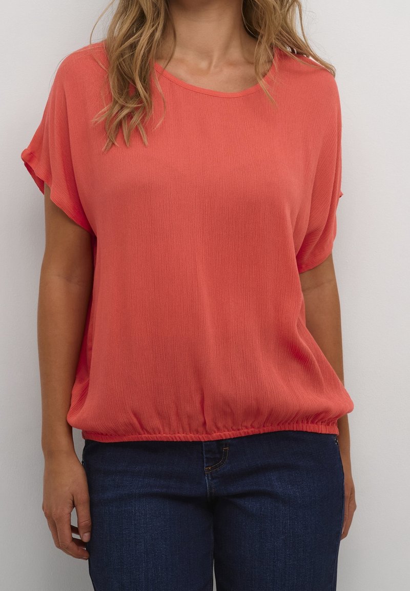 Femme portant un chemisier corail à manches courtes avec un ourlet élastique et un jean bleu foncé sur un fond uni.