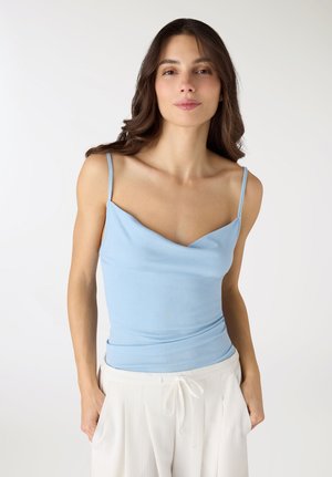 Donna con lunghi capelli scuri che indossa una canotta azzurra chiara e pantaloni bianchi con coulisse, in piedi davanti a uno sfondo bianco.