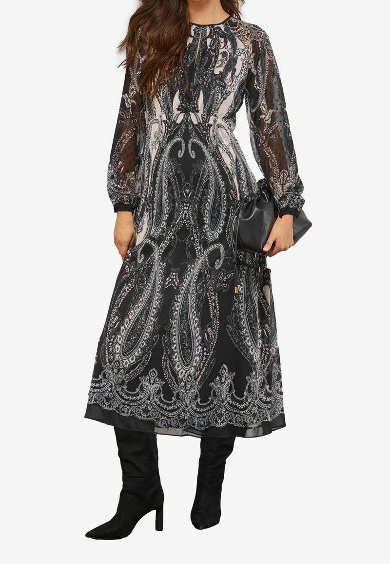 Robe midi à motif cachemire noir et blanc avec manches longues transparentes, associée à des bottes noires à talons et une pochette noire.