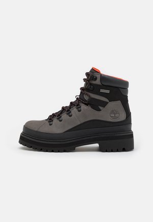 Botas de senderismo impermeables grises y negras con suela gruesa, detalles en rojo, cuello acolchado en el tobillo y cierre de cordones en la parte delantera sobre fondo blanco.