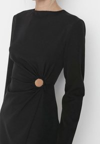 Robe noire en tissu lisse, dotée de manches longues, d'un col rond et d'une taille froncée avec un détail découpé circulaire.