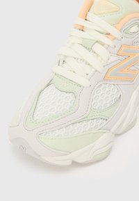 Sneaker vert clair et blanc avec des matériaux en mesh et en daim. Présente un grand logo orange "N", des lacets blancs et une semelle texturée.