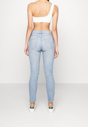 Vrouw draagt lichtblauwe hoge taille skinny jeans, witte asymmetrische cropped top met één schouder en witte Polo sneakers, staand met het gezicht naar een lichte muur gericht.