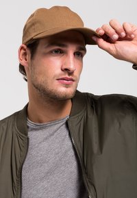 Khaki pet met een gebogen rand, schuin gedragen, gecombineerd met een olijfgroene bomberjack en een grijs T-shirt, wat een casual gelaagd uiterlijk laat zien.
