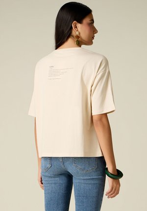 Kvinde med langt mørkt hår iført beige oversized T-shirt med tekst på bagsiden, blå jeans, stort grønt armbånd og hængende øreringe.