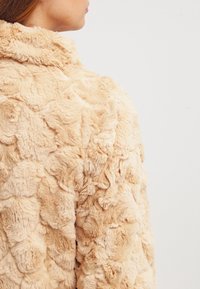 Faux fur jacka i ljusbeige med en texturerad, plyschig yta som har rundade, överlappande former över tyget.