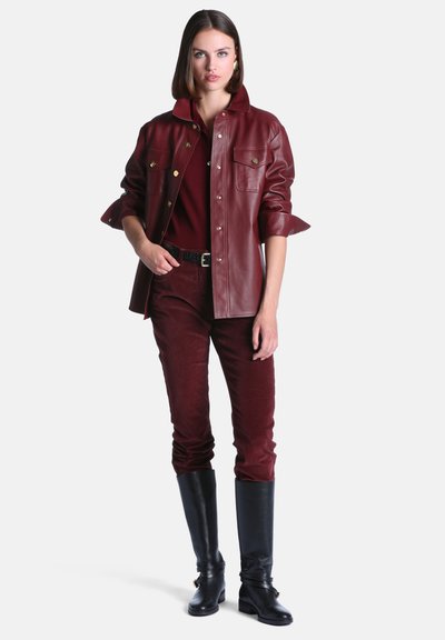 Femme portant une veste en cuir bordeaux, un pantalon assorti, une ceinture noire et de grandes bottes noires, debout avec une main dans la poche.