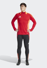 Rood en oranje sportshirt met lange mouwen, voorzien van een kwart-rits en zwarte sportleggings. Heeft gestikte accenten en een aansluitende pasvorm.