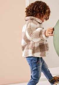 Tandy og hvidt ternet fleecejakke med krave, knaplukning og elastiske ærmer, iført over blå jeans og multicolor sko.