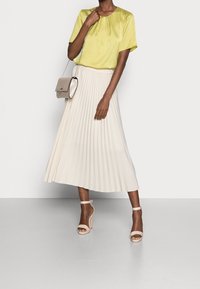 Blouse en satin vert clair avec un col rond, associée à une jupe plissée beige et des sandales à talons nus. Petit sac en bandoulière inclus.