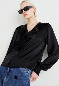 Blusa nera in satin con dettaglio arricciato sul davanti, scollatura a V e maniche lunghe. Abbinata a jeans in denim blu con un accento floreale nero.