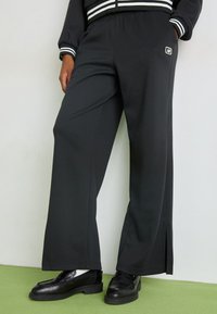 Pantaloni da jogging neri a gamba larga con vita elasticizzata, fessure laterali e una piccola patch del logo sulla tasca frontale. Tessuto testurizzato.