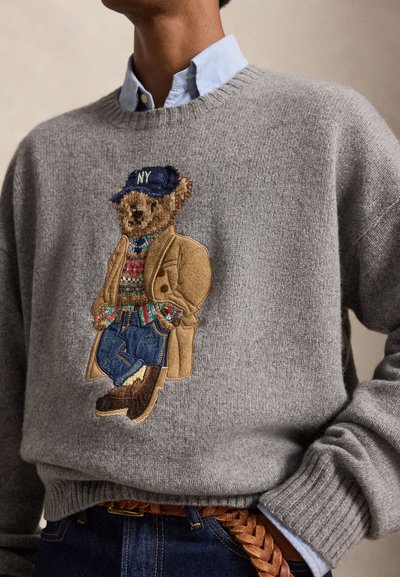 Personne portant un pull gris avec un ours brodé vêtu d'un manteau, d'un pull, d'un jean, de chaussures et d'une casquette marine avec l'inscription "NY".