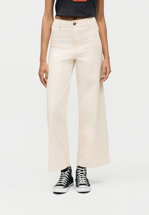 Naine, kes kannab beeže helesaarega laia lõikega kõrge vöökohaga pükse ja musti kõrge säärega tenniseid, seisab ühevärvilise hele tausta ees.