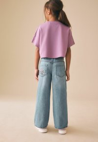 Copil cu păr lung, purtând un top scurt mov, blugi largi de culoare albastru deschis cu un patch brodat pe buzunarul din spate și adidași albi, stând cu fața spre spate.