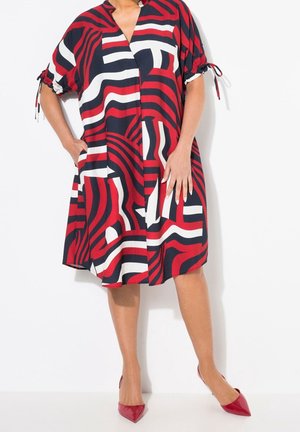 Czerwona i granatowa sukienka w wzory z dekoltem w serek, krótkimi rękawami i elastycznymi wiązaniami. Tkanina wydaje się lekka i zwiewna.