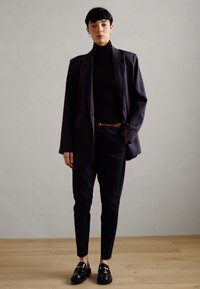 Marineblauer, übergroßer Blazer, eng anliegender schwarzer Rollkragenpullover, dunkle Hose und glänzende Slipper. Glatter Stoff mit minimaler Detailgestaltung.