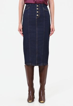 Donna con gonna di jeans blu scuro con bottoni frontali e stivali marroni fino al ginocchio, in piedi davanti a uno sfondo bianco uniforme.