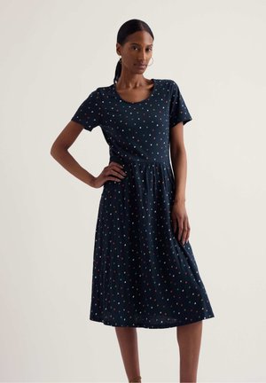 Kobieta ubrana w granatową sukienkę do kolan z krótkimi rękawami i wielokolorowymi kropkami, stojąca z jedną ręką na biodrze.