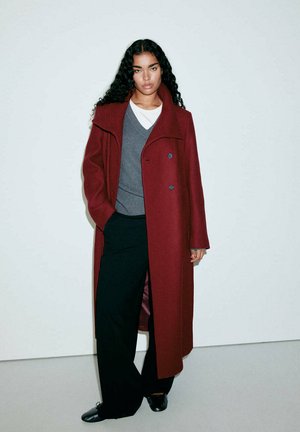 Femme aux longs cheveux bouclés portant un long manteau bordeaux, un pull gris, une chemise blanche, un pantalon noir et des chaussures noires, debout contre un mur uni.