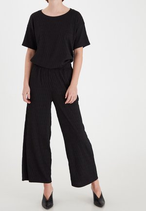 Persoon die een loszittend zwart getextureerd topje met korte mouwen en wijde broek met zwarte puntige schoenen met hak draagt tegen een witte achtergrond.