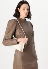 Rochie din piele maro cu mâneci lungi și umeri bufanți, combinată cu o geantă structurată de culoare crem, care prezintă un luciu subtil.