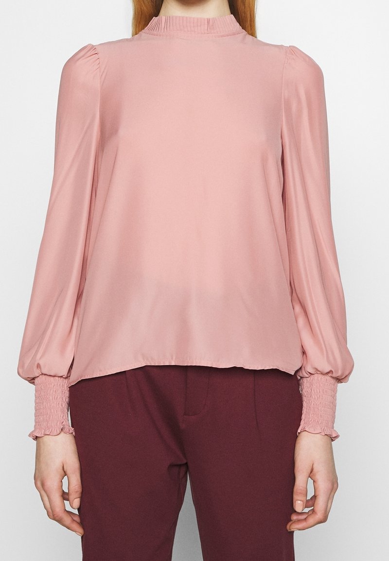 Personne portant un chemisier rose à manches longues avec des manches bouffantes et des poignets smockés, assorti à un pantalon bordeaux foncé.
