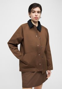 Γυναίκα που φορά καφέ μπουφάν με μαύρο γιακά και καφέ φούστα που ταιριάζει, στέκεται με το ένα χέρι στην τσέπη του μπουφάν μπροστά σε απλό φόντο.