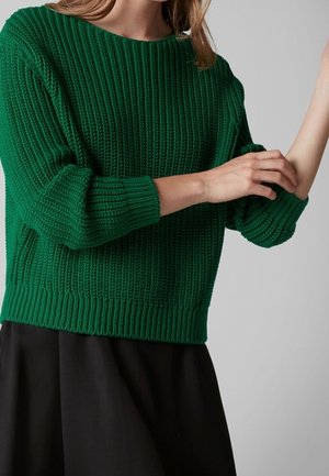 Kvinde justerer ærmet på en mørkegrøn strikket sweater, iført et sort skørt, mod en ensfarvet lys grå baggrund.