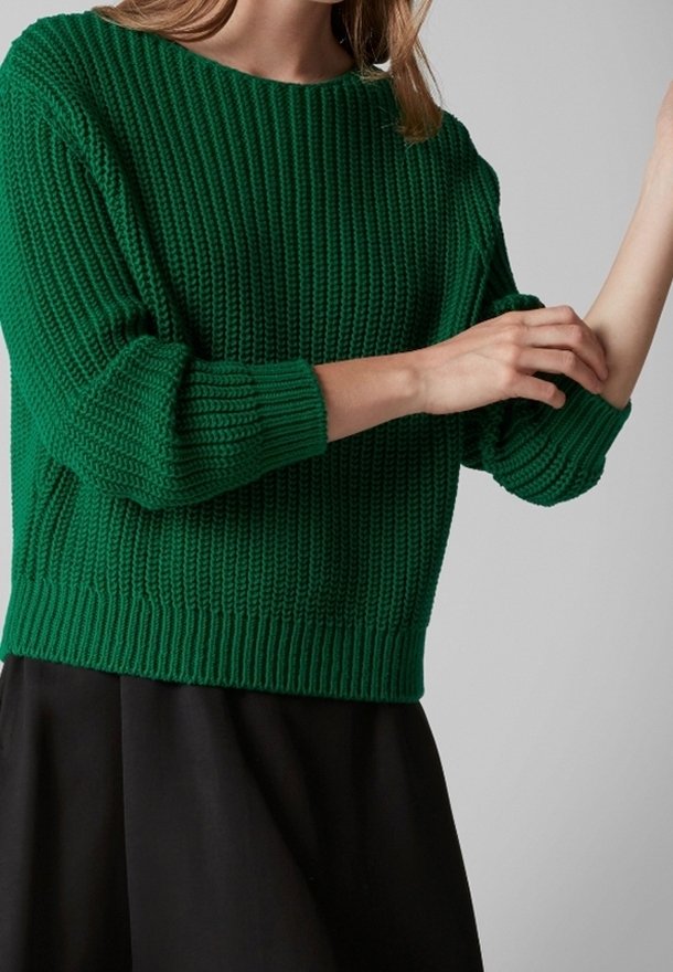 Femme ajustant la manche d'un pull en maille vert foncé, portant une jupe noire, sur un fond gris clair uni.