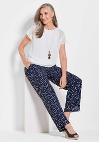 Weißes Kurzarm-Blusenshirt kombiniert mit einer dunkelblauen Hose mit Pünktchen. Glattes Material, lockere Passform. Die Schuhe haben ein Keilabsatzdesign.