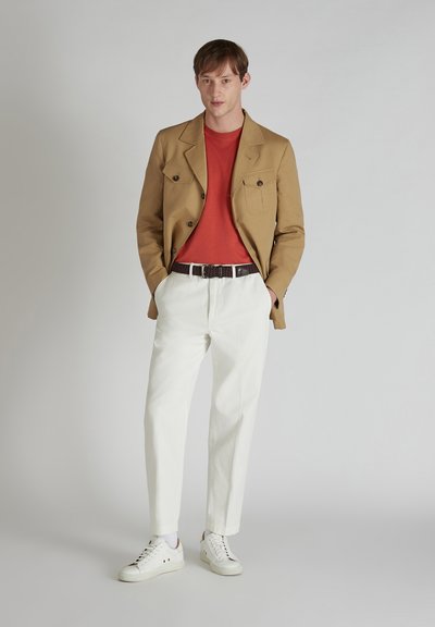 Beżowa marynarka dwurzędowa, czerwona koszulka z okrągłym dekoltem, białe spodnie, brązowy pasek i białe trampki. Gładkie tło.