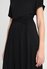 Musta mekko pehmeästä kankaasta, jossa on pyöreä pääntie, lyhyet hihat, solmittu vyötärökohta ja laskottu hameen design.