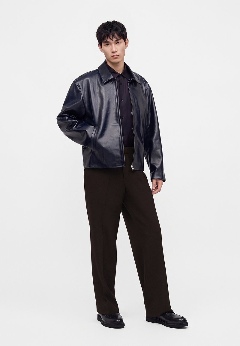 Joven con chaqueta de cuero negra, camiseta negra, pantalones marrón oscuro y zapatos negros, con una mano en el bolsillo de la chaqueta, sobre fondo blanco.