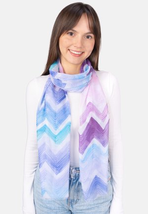 Junge Frau trägt einen pastellfarbenen Schal mit Chevron-Muster in Blau, Lila und Rosa über einem weißen Langarmshirt und hellblauen Jeans.