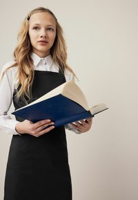 Meisje in een zwarte schort over een wit shirt, holding een groot blauw boek met een textuurcover. Zachte verlichting, neutrale achtergrond.