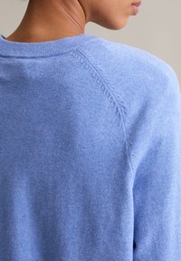 Posljednji kadar osobe koja nosi svijetloplavi pleteni pulover, s fokusom na detalje šava na ramenu i teksturiranu tkaninu.