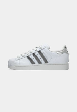 White Adidas Superstar sneaker with three black stripes, laces tied, rubber shell toe, and Adidas logo on the heel tab shown in side view.