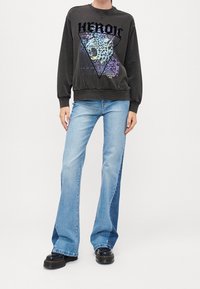 Persona che indossa un felpa nera con una grafica di un leone che ruggisce e il testo "HEROIC", jeans svasati blu chiaro e scarpe con plateau nere.