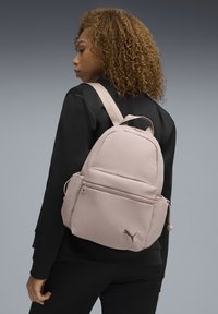 Sac à dos en tissu rose de forme arrondie, avec une poche avant zippée, des bretelles réglables et le logo Puma dans le coin inférieur. Texture lisse.