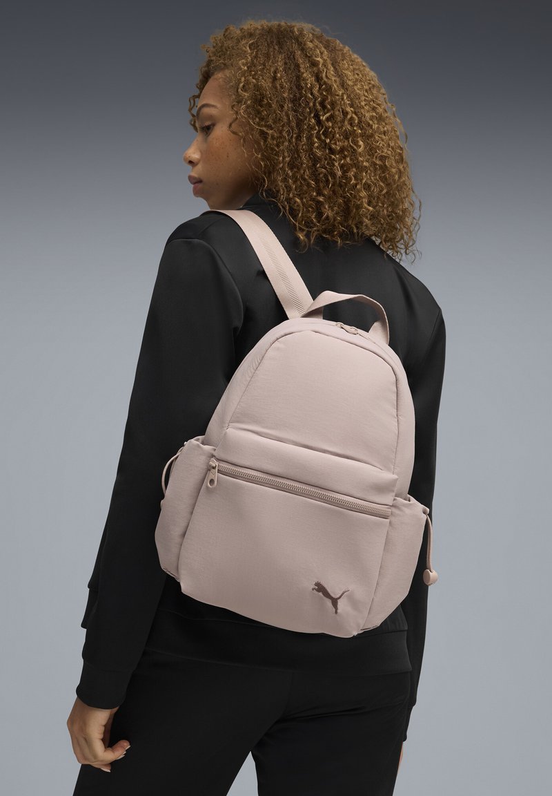 Sac à dos en tissu rose de forme arrondie, avec une poche avant zippée, des bretelles réglables et le logo Puma dans le coin inférieur. Texture lisse.