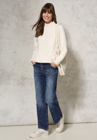 Suéter de punto blanco con un patrón texturizado, jeans de mezclilla azules y zapatillas blancas. Incluye un bolso cremoso y esponjoso colgado del hombro.