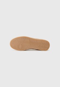 La semelle de chaussure présente une base en caoutchouc avec un motif de crampons en zigzag, un logo circulaire au centre, et une couleur marron clair avec des détails texturés.