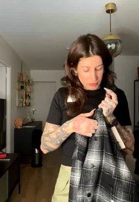 Frau mit Tattoos begutachtet einen schwarz-weiß karierten Mantel in einem modernen Wohnzimmer mit Holzboden und minimalistischer Einrichtung.