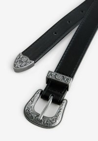 Ceinture en cuir noir avec une boucle en métal argenté ornée, présentant des motifs complexes et des pointes aux extrémités.