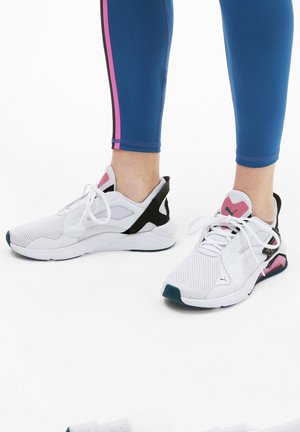 Baskets blanches de sport avec tige en mesh, accents noirs et roses, et bout arrondi, associées à des leggings bleus avec des rayures roses.