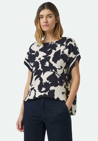 Kurzarmbluse in Marineblau und Creme mit einem floralen Muster. Lockere Passform, runder Halsausschnitt, leichte Stoffqualität. Kombiniert mit dunklen Jeans.