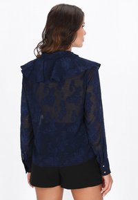Blusa azul marina translúcida con bordado floral, cuello con volantes y mangas largas. Puños con botones y textura suave. Llevada sobre pantalones cortos negros.