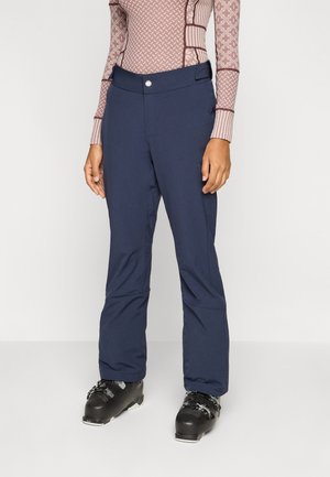 Person trägt marineblaue Skihose, schwarze Skischuhe und ein langärmeliges, gemustertes Oberteil vor einem einfarbigen hellen Hintergrund.