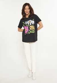 Sort bomulds T-shirt med farverigt blomster grafikdesign med pink, hvide og gule blomster samt grønne blade, parret med hvide bukser og sneakers.